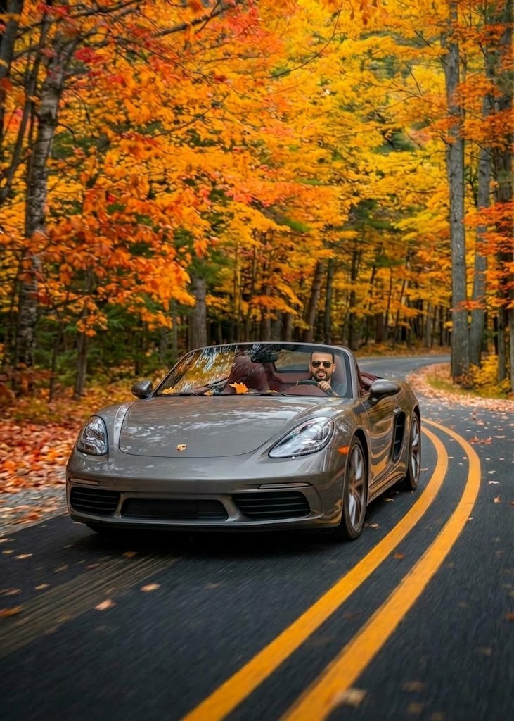 A silver convertible with its top down and a driver in sunglasses rounds a bend on a road lined with brilliant autumn leaves, double yellow lines curving ahead.